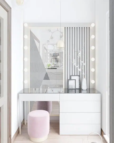White makeup vanity with a glass top and three drawers, surrounded by round bulbs, a pale pink upholstered stool, a large illuminated mirror, decorative vases and frames on the surface, and gray curtains to the right.
